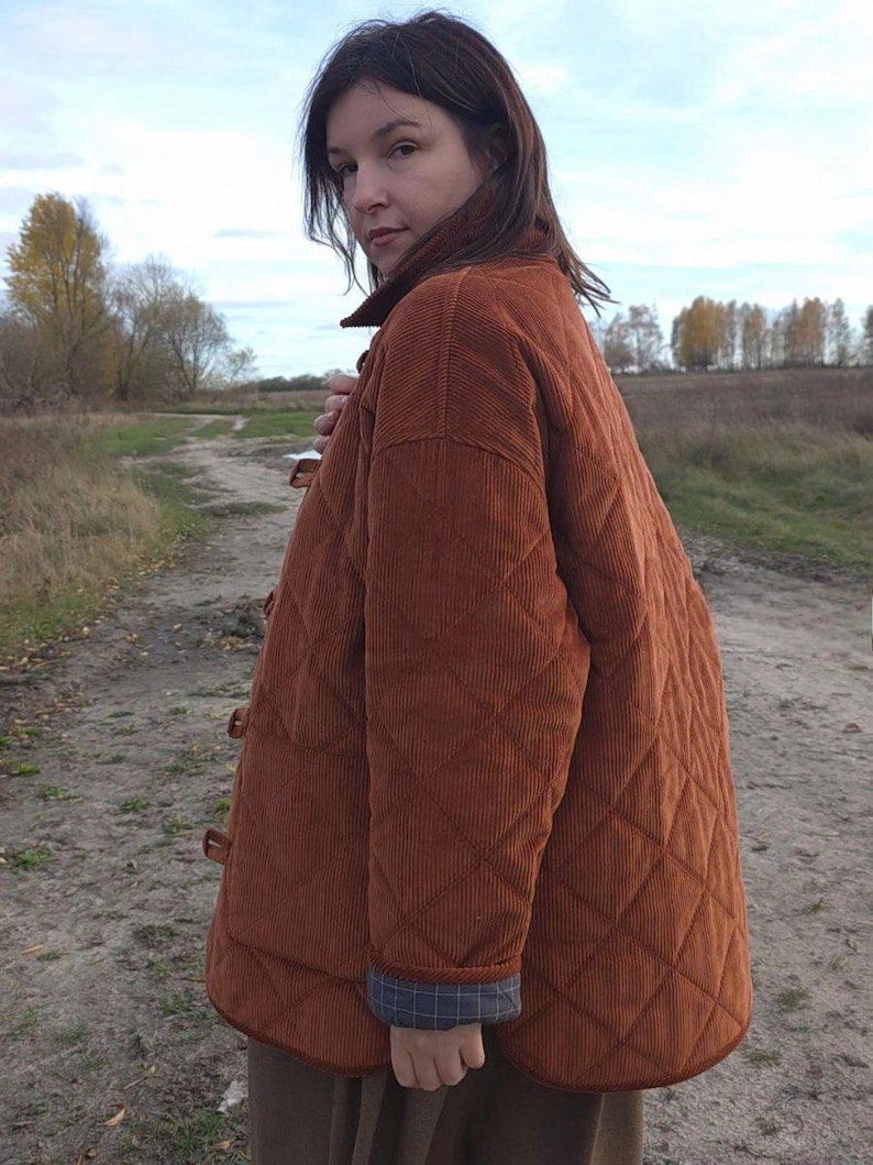 May include: A quilted, rust-colored corduroy jacket with a diamond pattern. The jacket has a button-up front and a plaid lining at the cuffs. The person is standing on a dirt path in a field.