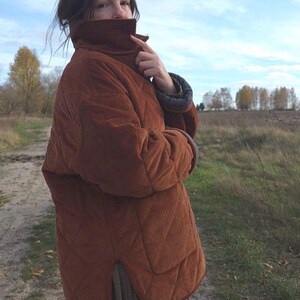 May include: A quilted, rust-colored corduroy jacket with a high collar. The jacket has a diamond pattern and a relaxed fit. The sleeves have a contrasting blue and gray lining. The person is standing outdoors in a field.