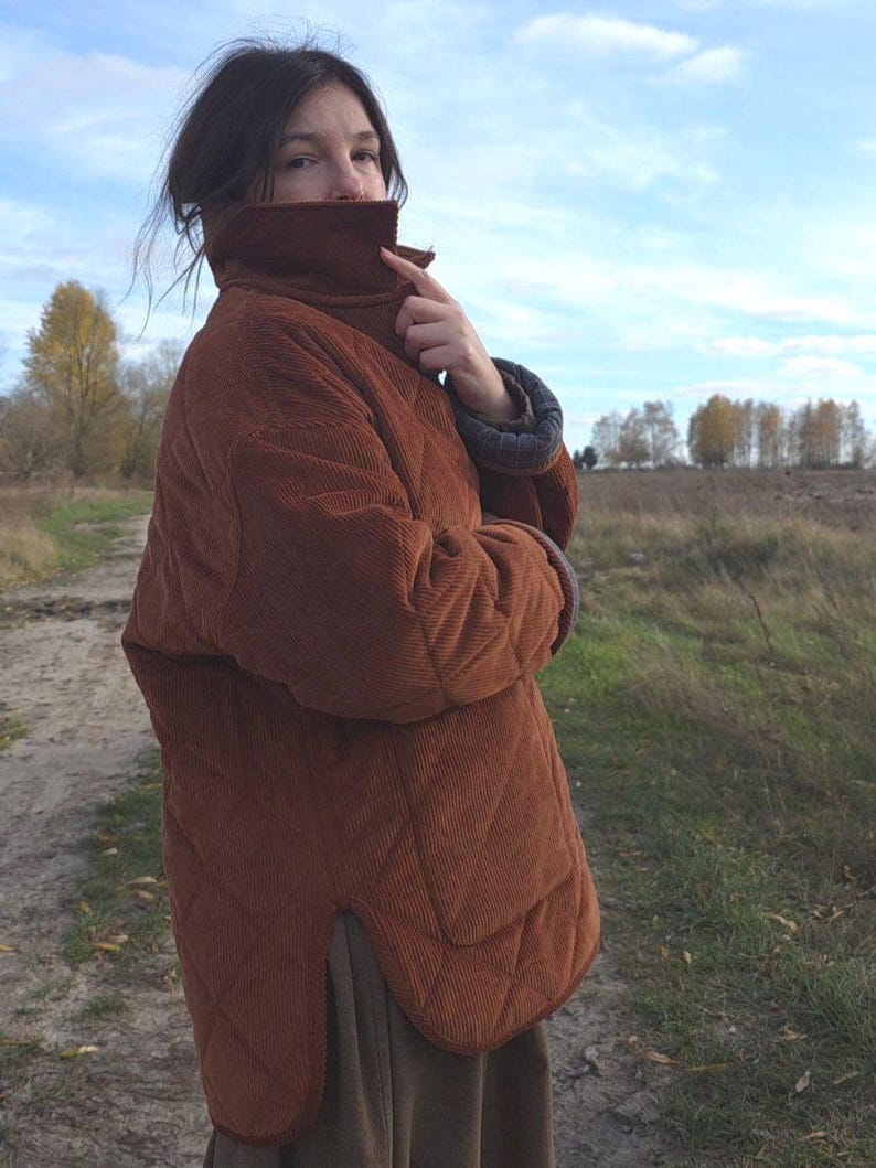 May include: A quilted, rust-colored corduroy jacket with a high collar. The jacket has a diamond pattern and a relaxed fit. The sleeves have a contrasting blue and gray lining. The person is standing outdoors in a field.