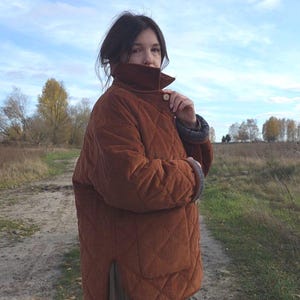May include: A quilted, corduroy jacket in a warm, reddish-brown hue. The jacket features a high collar, button closure, and a diamond pattern. The sleeves have a contrasting lining. The jacket is worn outdoors on a cloudy day.