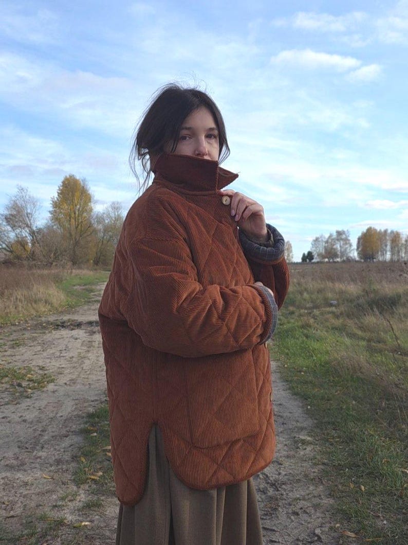 May include: A quilted, corduroy jacket in a warm, reddish-brown hue. The jacket features a high collar, button closure, and a diamond pattern. The sleeves have a contrasting lining. The jacket is worn outdoors on a cloudy day.