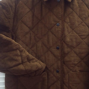 May include: A brown corduroy jacket with a quilted diamond pattern. The jacket has a collar, black buttons, and two front pockets. The sleeves are long, and the jacket appears to be made of a soft, textured fabric.