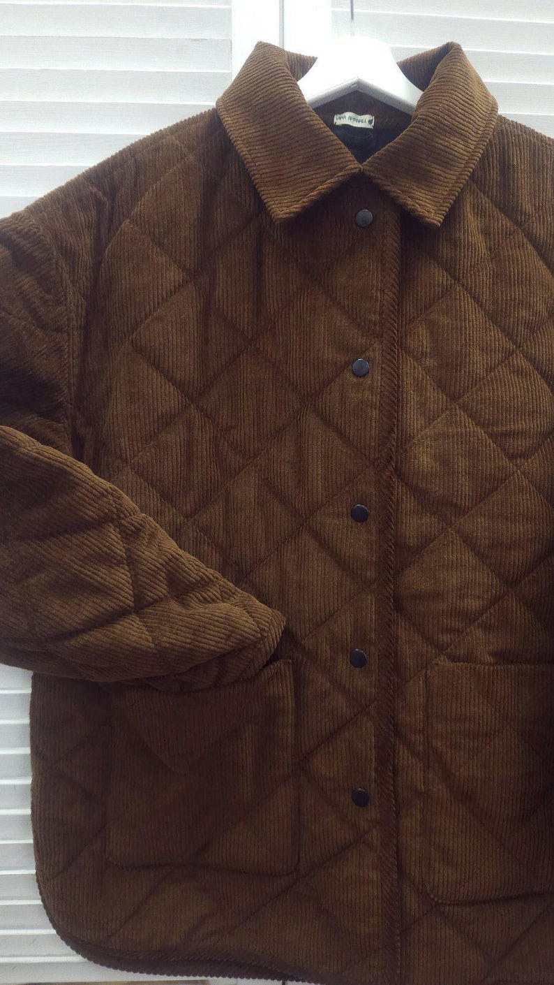 May include: A brown corduroy jacket with a quilted diamond pattern. The jacket has a collar, black buttons, and two front pockets. The sleeves are long, and the jacket appears to be made of a soft, textured fabric.