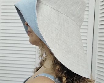 May include: A wide-brimmed bucket hat featuring a reversible design with a natural beige linen exterior and a contrasting light blue interior lining. The hat has a structured crown and a downward-sloping brim that provides coverage around the head.