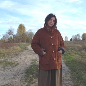 May include: A rust-colored quilted jacket with a high collar and wooden toggle closures. The jacket has pockets and is worn over a long, olive-green skirt. The outfit is worn outdoors on a dirt path with a field and trees in the background.