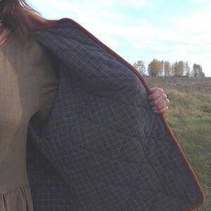 May include: A brown quilted jacket with a dark gray checkered lining. The jacket has a brown corded trim and is open, revealing the interior. The background shows a field and trees under a blue sky.