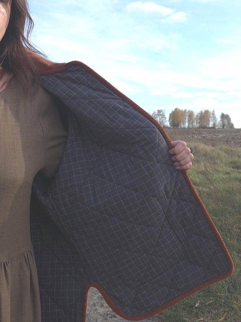 May include: A brown quilted jacket with a dark gray checkered lining. The jacket has a brown corded trim and is open, revealing the interior. The background shows a field and trees under a blue sky.