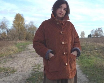 May include: A rust-colored quilted jacket featuring a high collar, long sleeves with grey patterned cuffs, and a front closure with four light-colored toggle buttons. The jacket includes two large patch pockets on the front and a rounded hemline.