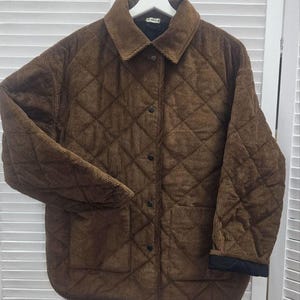 May include: A brown corduroy jacket with a quilted diamond pattern. The jacket has a collar, button closure, and two front pockets. The cuffs are black. The jacket is hanging on a white hanger.