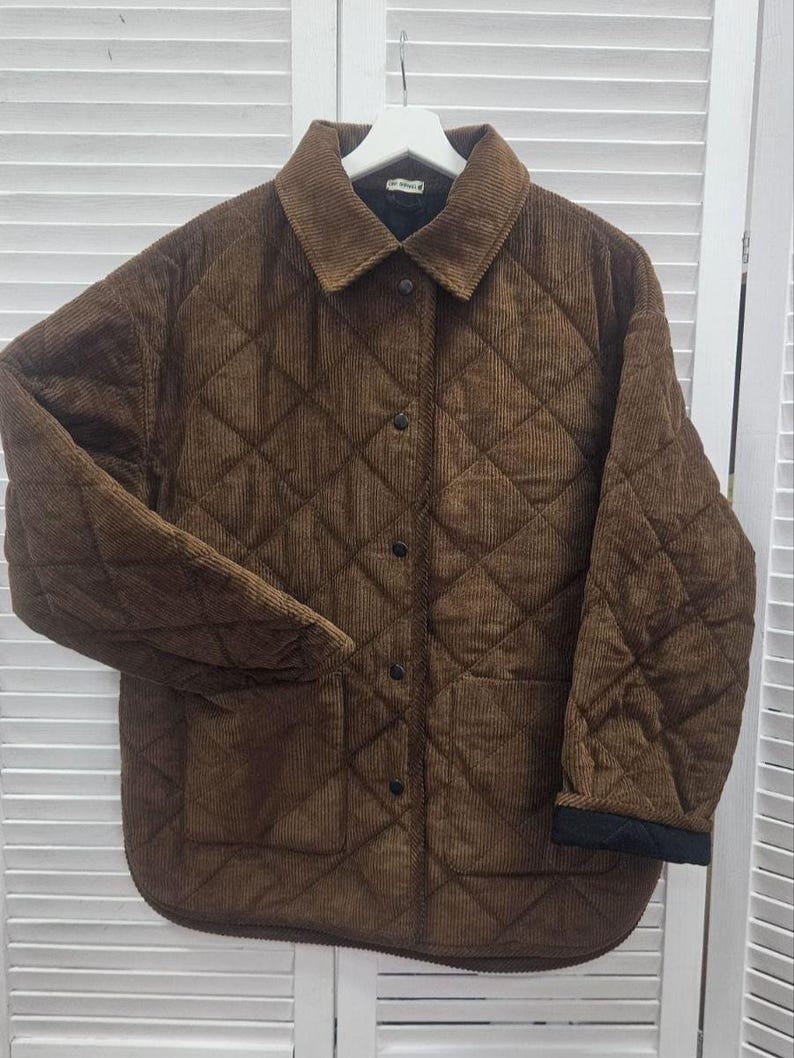 May include: A brown corduroy jacket with a quilted diamond pattern. The jacket has a collar, button closure, and two front pockets. The cuffs are black. The jacket is hanging on a white hanger.