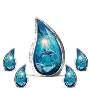 May include: A set of silver-coloured, teardrop-shaped pendants and earrings. Each piece features a blue ocean scene with a dolphin and a bright sun. The jewellery has a detailed, textured border.
