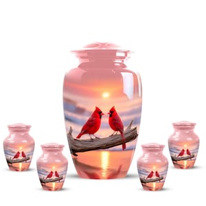 May include: Pink cremation urns with a sunset scene and red cardinal birds perched on a branch. The urns are in various sizes, with a large urn and several smaller keepsake urns.