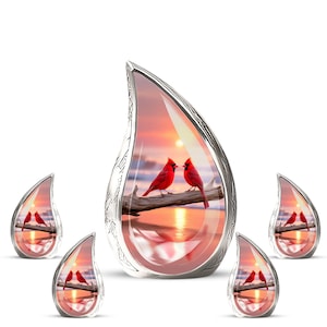 May include: A collection of teardrop-shaped pendants with a silver-colored trim. Each pendant features a sunset scene with two red cardinals perched on a branch. The background shows a sunset over water.