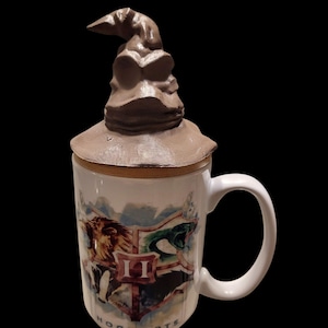 May include: White ceramic mug with a brown Sorting Hat lid. The mug features the Hogwarts crest with a lion, snake, eagle, and badger. The word "HOGWARTS" is printed at the bottom of the mug.