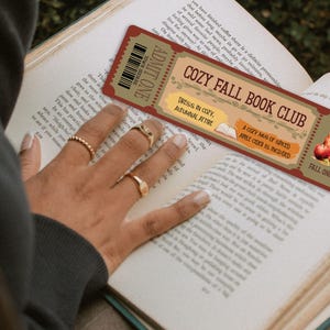 May include: A red and brown bookmark with the text "Cozy Fall Book Club" and a picture of a mug of apple cider. The bookmark also says "Dress in cozy, autumnal attire" and "A cozy mug of spiced apple cider is included".