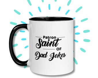 May include: White ceramic mug with a black interior and handle. The mug features the text "Patron Saint of Dad Jokes" in a black, stylized font. The design is set against a white background with blue accents.