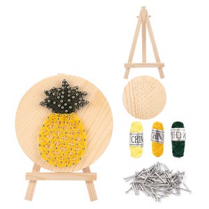 Op de afbeelding: Een doe-het-zelf string art kit met een ananas ontwerp. De kit bevat een houten basis, spijkers, geel, groen en zwart garen, en een kleine houten ezel. De ananas is gemaakt met gele draad en heeft een groene top.