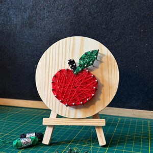 May include: A handmade string art apple on a wooden circle, mounted on a small easel. The red apple is crafted with string and surrounded by silver pins. A green leaf and black stem complete the design. Craft supplies are visible nearby.
