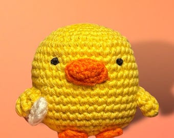 Puede incluir: Un juguete de pollito amarillo tejido a crochet con pico y patas naranjas. El pollito sostiene un objeto blanco tejido a crochet en su ala derecha.