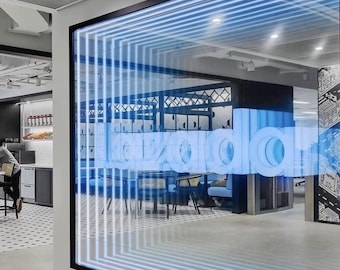 May include: A large glass partition featuring the word Lazada in bold, blue, three-dimensional lettering. The glass is framed in black and decorated with a series of repeating, glowing blue horizontal lines that create a tunnel-like visual effect.