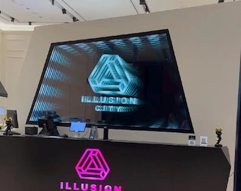 May include: A black reception desk featuring a glowing pink logo and the text "ILLUSION CITY". Above the desk, a large trapezoidal screen displays a repeating, three-dimensional geometric triangle logo with the text "ILLUSION CITY" in white.