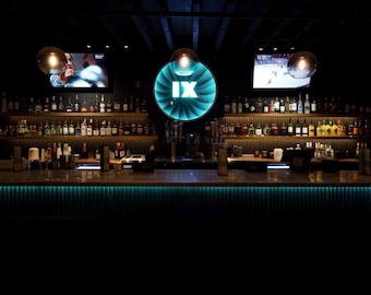 May include: A dimly lit bar interior featuring a long counter with teal under-lighting. Behind the bar, shelves hold numerous glass bottles. A circular sign with the Roman numeral IX glows in teal, flanked by two television screens showing sports.