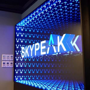 Op de afbeelding: Een verlicht bord met het woord "SKYPEAK" in witte, 3D-letters. De achtergrond is een raster van blauwe lichten, met vraagtekens bovenaan. Het bord is bevestigd aan een donkere muur.