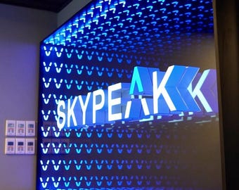 May include: A large, illuminated sign with the word "SKYPEAK" in white, 3D block letters. The background is a grid of blue lights, with a pattern of question marks at the top. The sign is mounted on a dark wall.