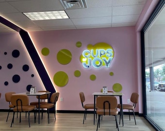 May include: A cafe interior featuring a light pink wall with a large, illuminated sign reading CUPS JOY. The wall is decorated with lime green circles and a dark purple diagonal accent. Two square tables with tan chairs are arranged on light wood flooring.
