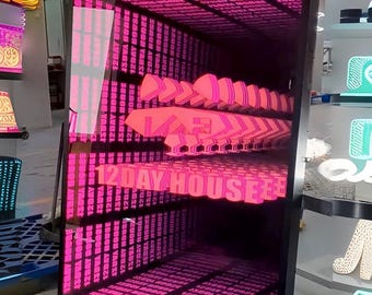 May include: A rectangular infinity mirror sign with a black frame featuring the text 12 DAY HOUSE in bright pink neon lettering. The internal lighting creates a repeating, tunnel-like visual effect of the text receding into the distance.