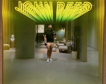 May include: A square-framed mirror installation featuring a repeating, infinite tunnel effect. The bright yellow neon text reads JOHN REED, which is reflected multiple times, creating a deep, glowing visual corridor within the reflective glass surface.
