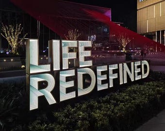 May include: A large, illuminated outdoor sign featuring the words LIFE REDEFINED in block letters. Each letter displays an internal infinity mirror effect with repeating white light patterns, set against a dark structure at night.