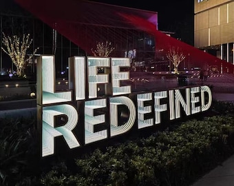 May include: A large, illuminated outdoor sign featuring the words LIFE REDEFINED in block letters. Each letter displays an internal infinity mirror effect with repeating white light patterns, creating a deep, tunnel-like visual illusion.