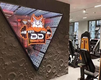 May include: A triangular infinity mirror wall sign featuring an orange and white monster mascot holding a barbell with the text DD MONSTER. The sign is mounted on a dark gray textured wall with crater-like patterns inside a gym with exercise equipment.