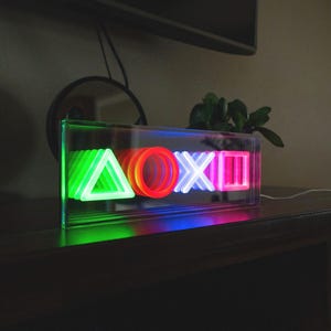 May include: A clear acrylic box with neon lights shaped like the PlayStation controller symbols: a green triangle, a red circle, a blue X, and a pink square.