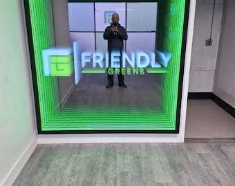 May include: A square wall-mounted infinity mirror featuring a black frame and bright green LED lighting that creates a tunnel effect. The center of the mirror displays the white text Friendly Greens with a stylized green logo to the left side.