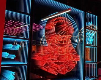 May include: A wall-mounted neon light installation featuring a bright red astronaut figure centered within a series of concentric blue rectangular frames that create an infinite tunnel effect. The display is positioned above a white dining table.