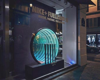 May include: A storefront display featuring a circular infinity mirror with teal lighting and five vertical black bars. The wall displays the text NIKE+ FUELBAND SE and MOTIVÉ. PARTOUT. TOUT LE TEMPS. The base includes the text JUST DO IT. and a Nike logo.