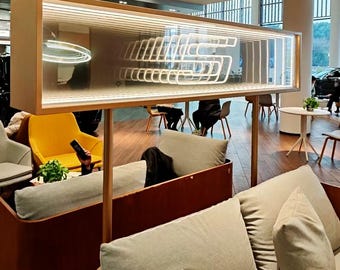 May include: A rectangular infinity mirror display featuring a glowing white neon letter S, mounted on two thin metal poles above a seating area with light gray cushions and a yellow armchair in a modern interior space with wooden flooring.
