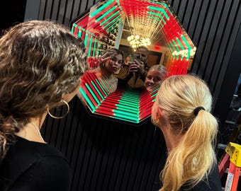 May include: An octagonal infinity mirror with red and green LED lights reflecting a group of people. The mirror is mounted on a black wall, and two people are looking at the mirror.