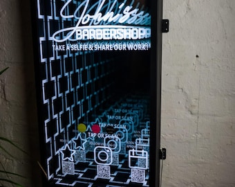 May include: A rectangular black wall-mounted infinity mirror sign for John's Barbershop. The display features white neon-style text reading John's Barbershop and Take a Selfie & Share Our Work, with QR codes, star, camera, and calendar icons.