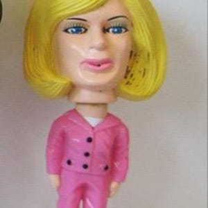 May include: A bobblehead doll featuring a woman with yellow hair, blue eyes, and pink lips. She wears a pink suit with black buttons and black shoes. The doll's head is disproportionately large compared to the body.