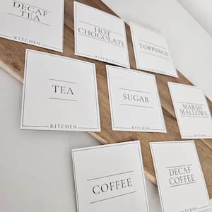 K&ouml;nnte beinhalten: Wei&szlig;e quadratische Etiketten mit schwarzem Text f&uuml;r eine Hei&szlig;getr&auml;nkebar. Die Etiketten lauten "Kaffee ohne Koffein", "Hei&szlig;e Schokolade", "Toppings", "Tee", "Zucker", "Marshmallows", "Kaffee" und "Kaffee ohne Koffein". Jedes Etikett tr&auml;gt den Schriftzug "K&uuml;che" am unteren Rand.