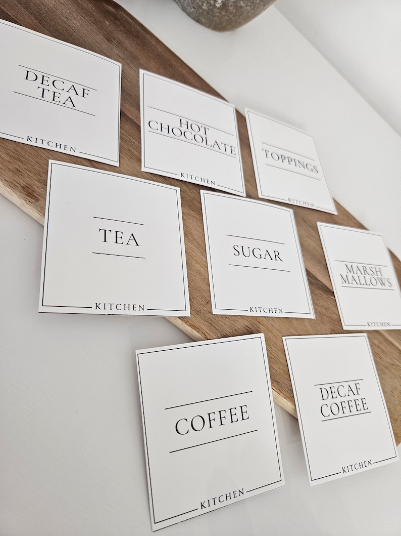 K&ouml;nnte beinhalten: Wei&szlig;e quadratische Etiketten mit schwarzem Text f&uuml;r eine Hei&szlig;getr&auml;nkebar. Die Etiketten lauten "Kaffee ohne Koffein", "Hei&szlig;e Schokolade", "Toppings", "Tee", "Zucker", "Marshmallows", "Kaffee" und "Kaffee ohne Koffein". Jedes Etikett tr&auml;gt den Schriftzug "K&uuml;che" am unteren Rand.