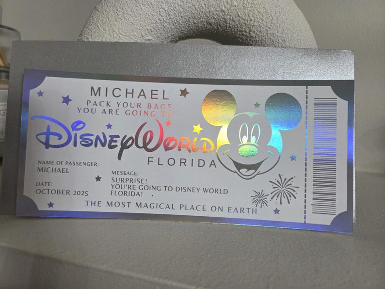 May include: A silver ticket-style announcement for a trip to Disney World in Florida. The ticket features the Disney World logo, a Mickey Mouse graphic, and the name "MICHAEL." It includes the message "PACK YOUR BAGS YOU ARE GOING TO..." and "THE MOST MAGICAL PLACE ON EARTH."