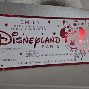 May include: A red and white Disneyland Paris ticket with the name "EMILY" and the date "OCTOBER 2025". The ticket features the Disneyland logo and a Minnie Mouse illustration. The text reads "PACK YOUR BAGS, YOU ARE GOING TO..."