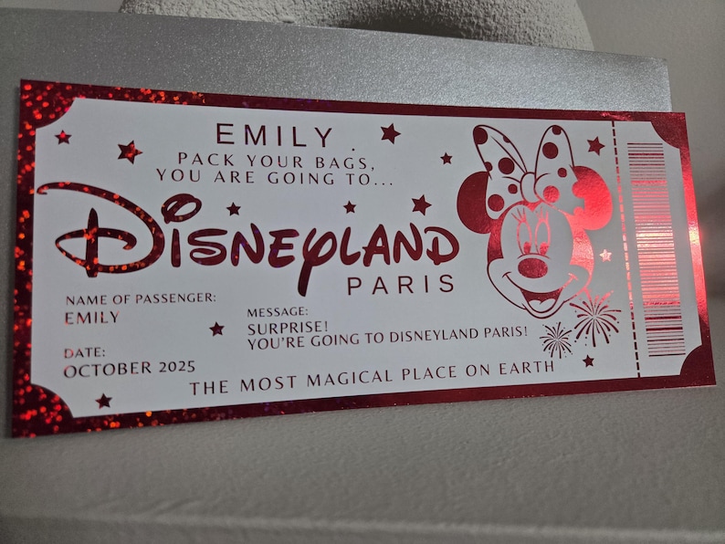 May include: A red and white Disneyland Paris ticket with the name "EMILY" and the date "OCTOBER 2025". The ticket features the Disneyland logo and a Minnie Mouse illustration. The text reads "PACK YOUR BAGS, YOU ARE GOING TO..."