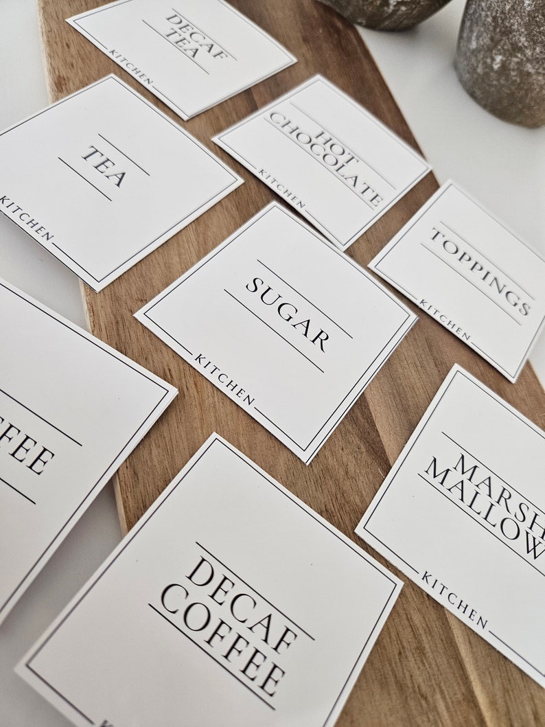 K&ouml;nnte beinhalten: Wei&szlig;e quadratische Etiketten mit schwarzem Text f&uuml;r eine hei&szlig;e Getr&auml;nkebar. Die Etiketten lauten "Kaffee ohne Koffein", "Hei&szlig;e Schokolade", "Toppings", "Tee", "Zucker", "Marshmallow", "Kaffee" und "Kaffee ohne Koffein".