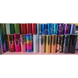 May include: A collection of colorful vinyl rolls in various shades, including green, red, blue, and gold. Some rolls have glitter or holographic effects. The rolls are arranged in rows, showcasing a variety of colors and textures.