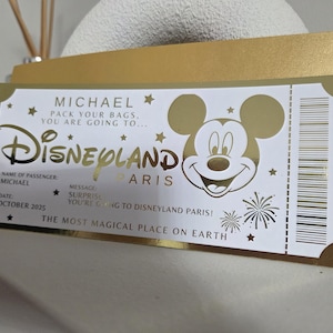 May include: A gold and white Disneyland Paris ticket-themed card. The card features the Disneyland logo, Mickey Mouse, and text that reads "MICHAEL, PACK YOUR BAGS, YOU ARE GOING TO..." and "THE MOST MAGICAL PLACE ON EARTH."